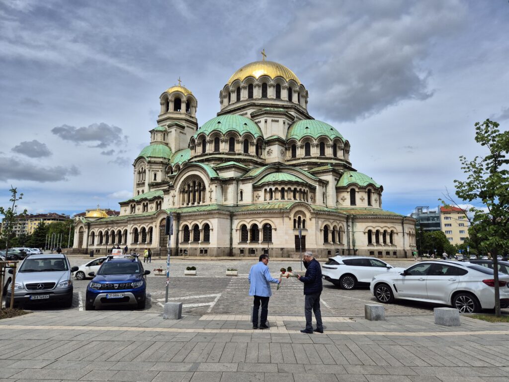 Zdjęcie prezentuje monumentalny sobór św. Aleksandra Newskiego w Sofii, którzy stanowi tło sceny przedstawiającą mężczyzn przekazujących sobie kwiaty.