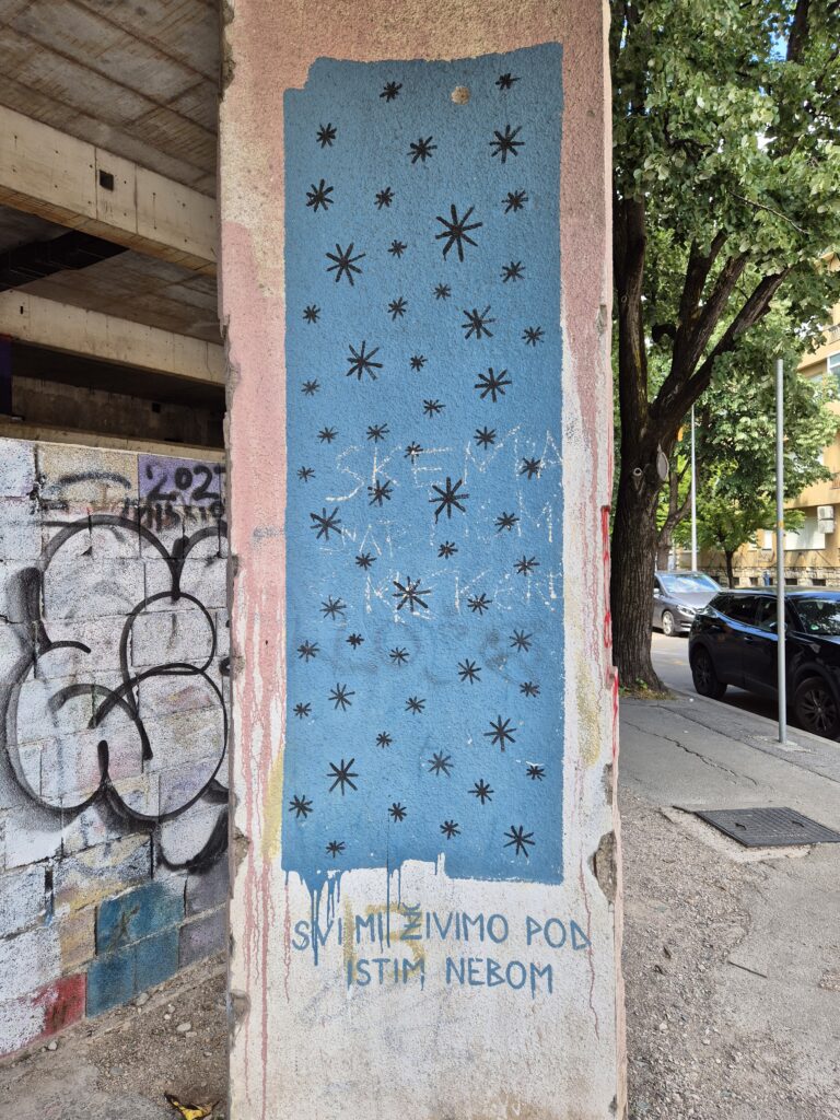 Zdjęcie przedstawia mural z napisem "Svi mi živimo pod istim nebom".